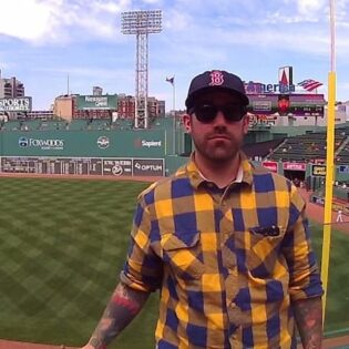 Paul Ledoux donning a Red Sox baseball cap standing in Fenway Park bleachers across the field from the Green Monster